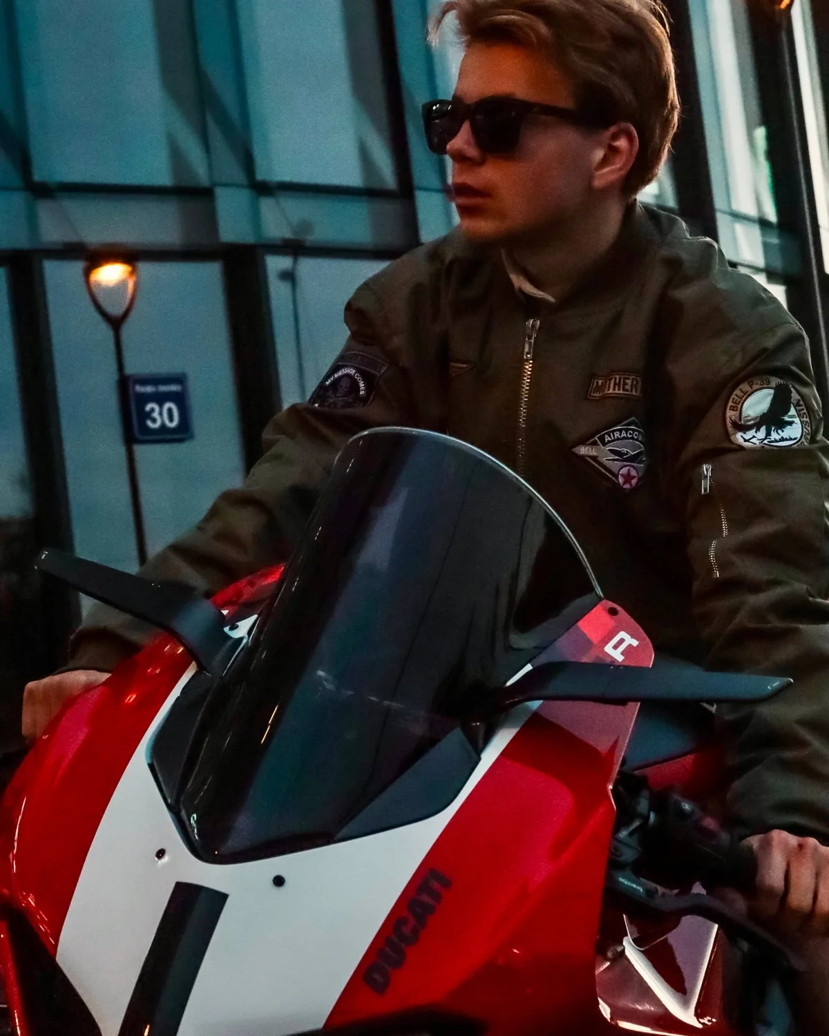 Aleksandrs on a red sport motorcycle at dusk, sunglasses and a bomber jacket